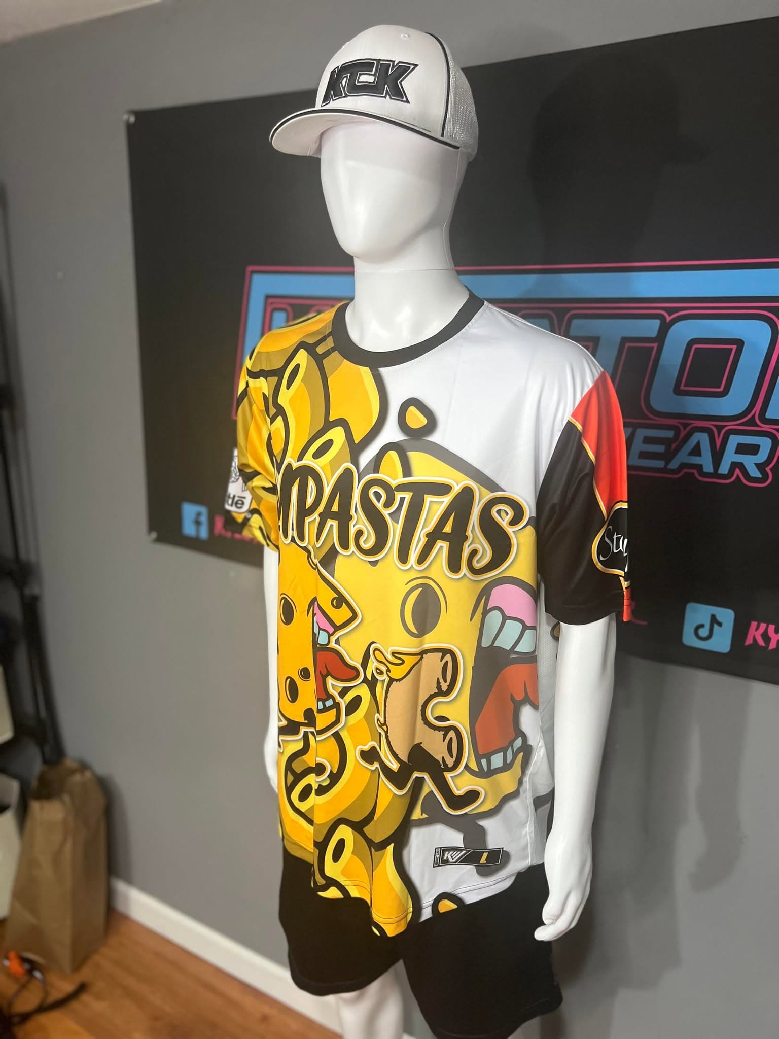 The first Impastas jersey on a mannequin at the print shop: white with a large yellow cheese-and-noodle cartoon across the front, IMPASTAS in black-outlined letters, a black collar and a red and black sleeve