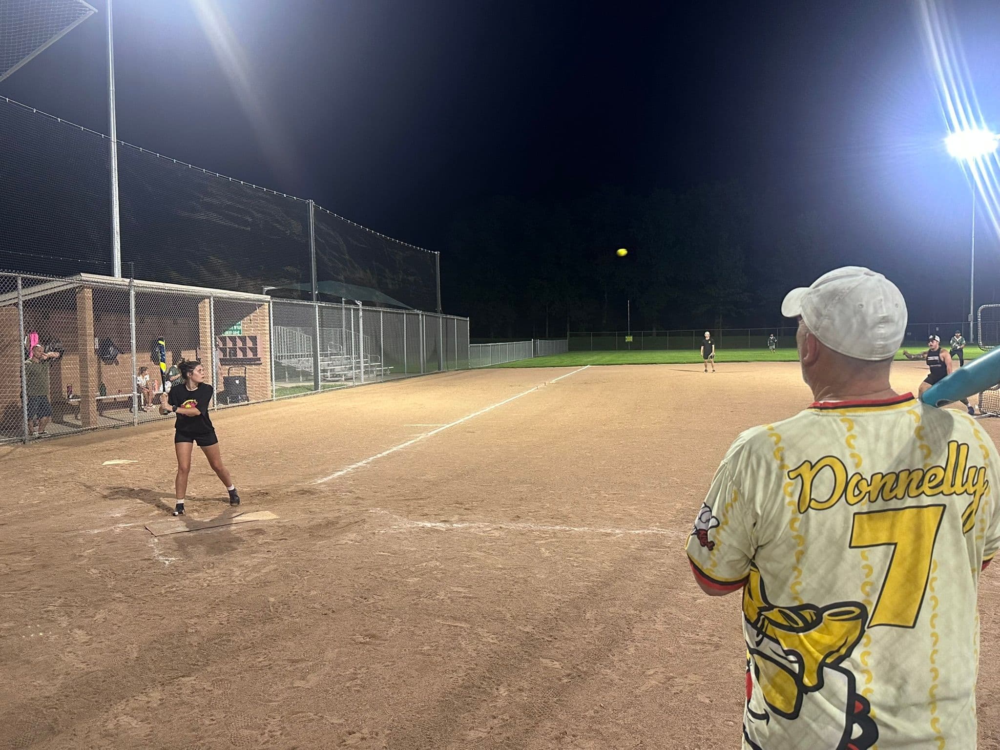 Emma Musick out on the infield dirt as the pitch goes in, seen past Scott Donnelly's number 7 Impastas jersey in the foreground, under the lights