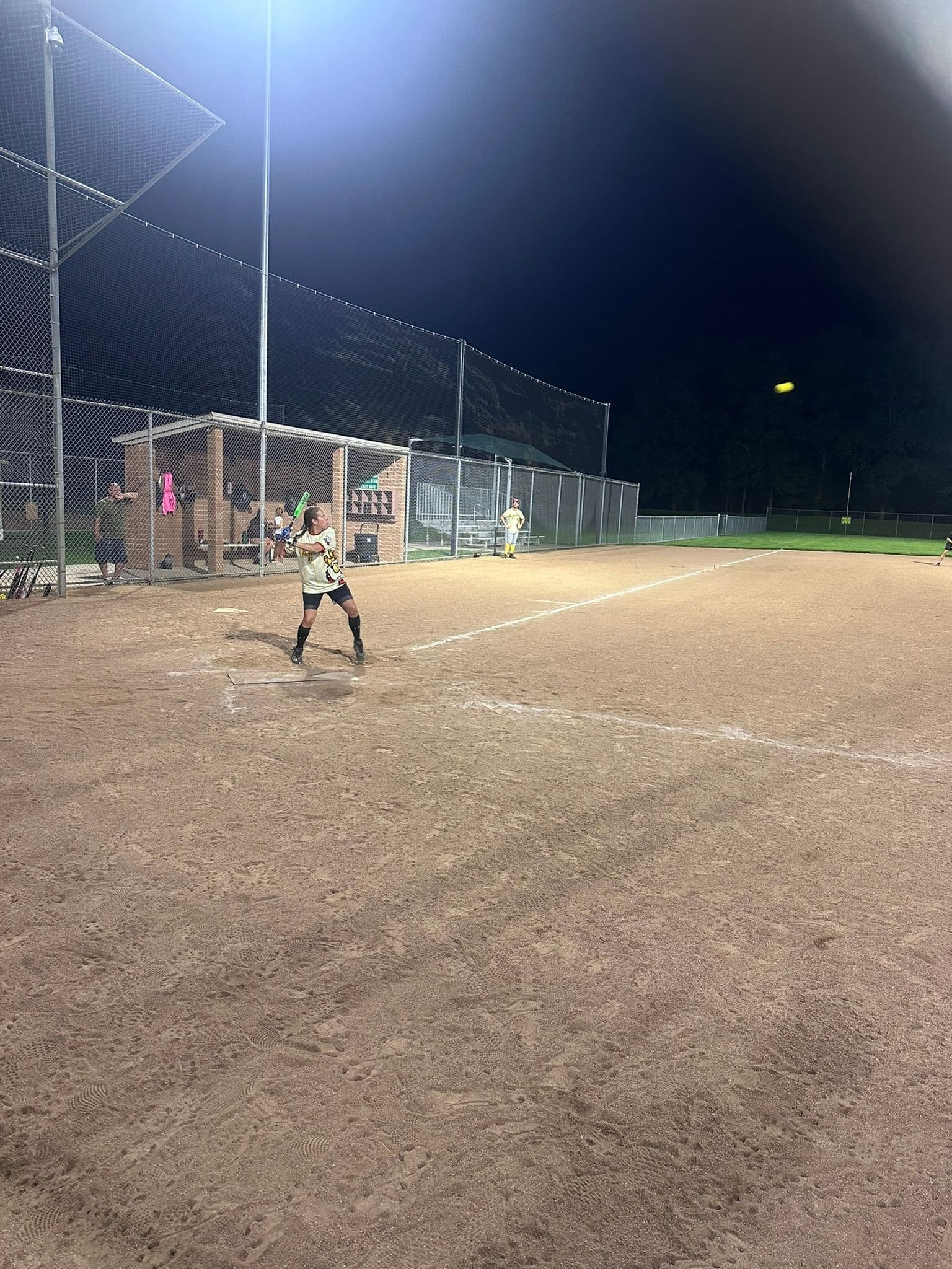 Wide view of the whole field at night from behind home plate, Jacki White standing in at the plate, the dugout and backstop down the line and the ball in flight