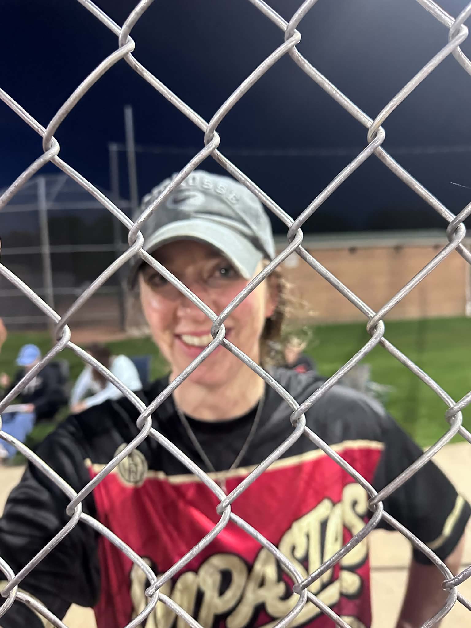 Isabel Simon smiling through the chain-link backstop in her Impastas jersey and a pale cap, the bench and the lit outfield behind her