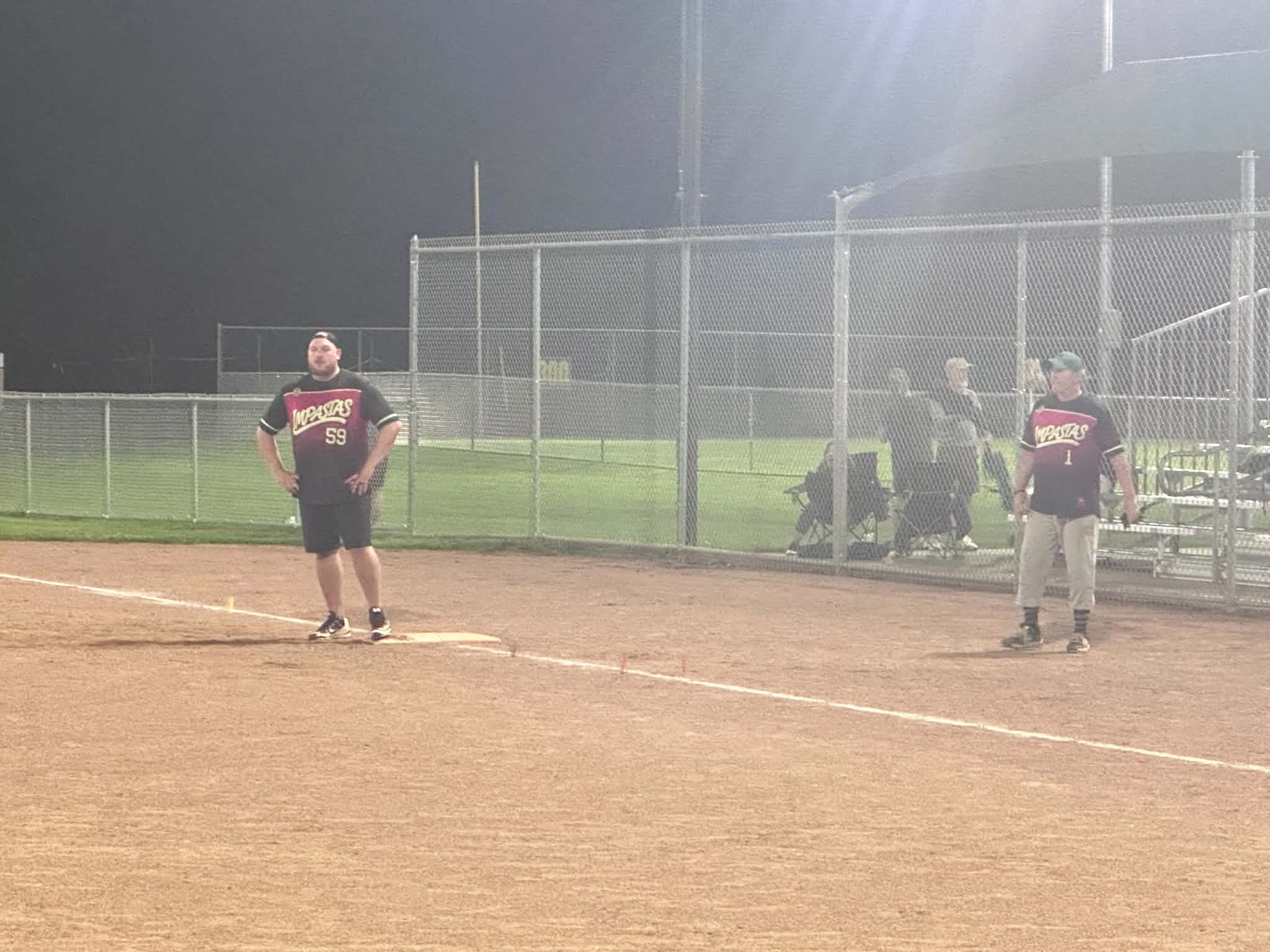 Adam Highberger standing on the base in number 59 and Scott Donnelly down the line in number 1, both in the new Impastas jerseys, spectators in camp chairs beyond the fence